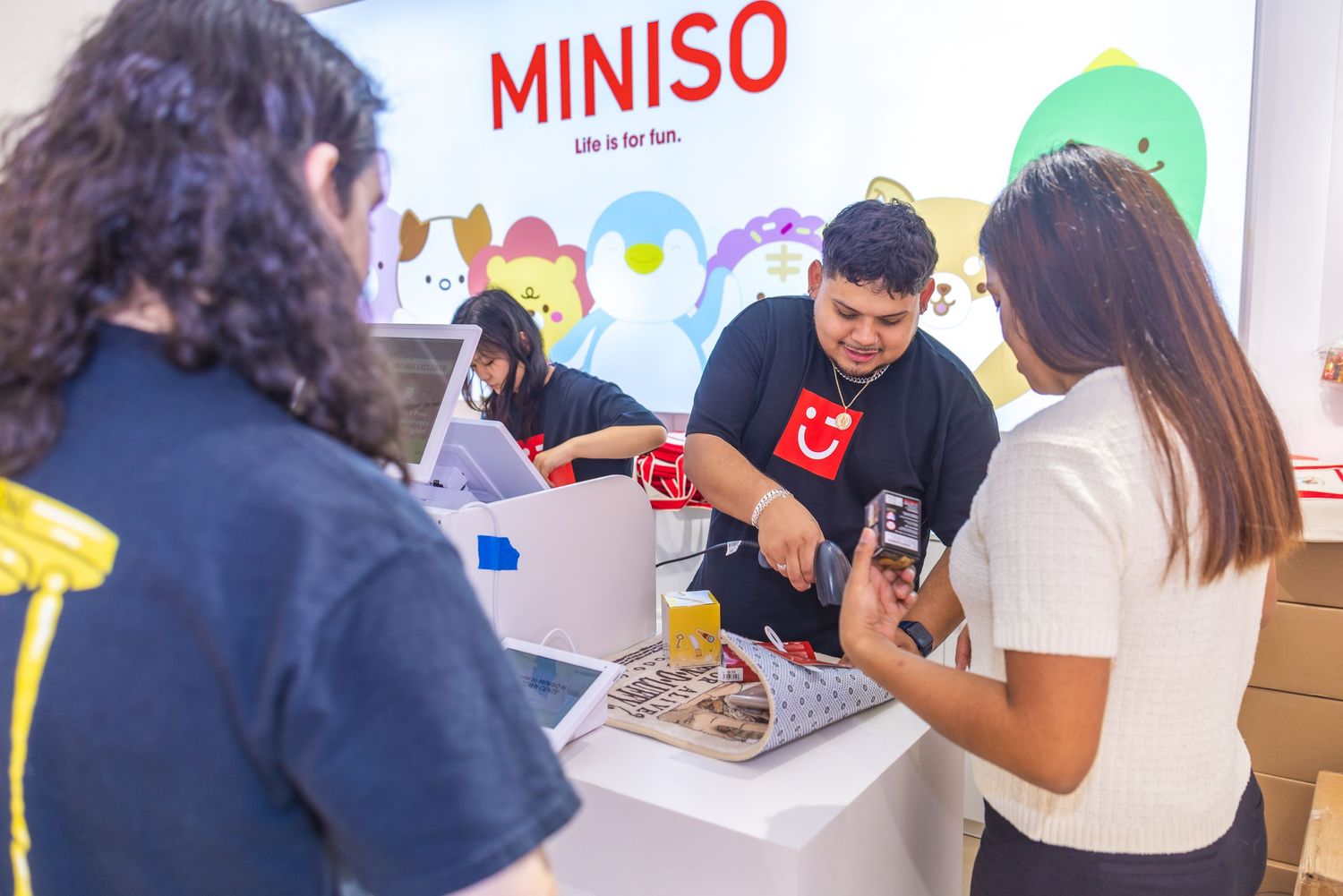 Miniso staff member assisting customers at a product demonstration table inside a bright store with the brand logo displayed.