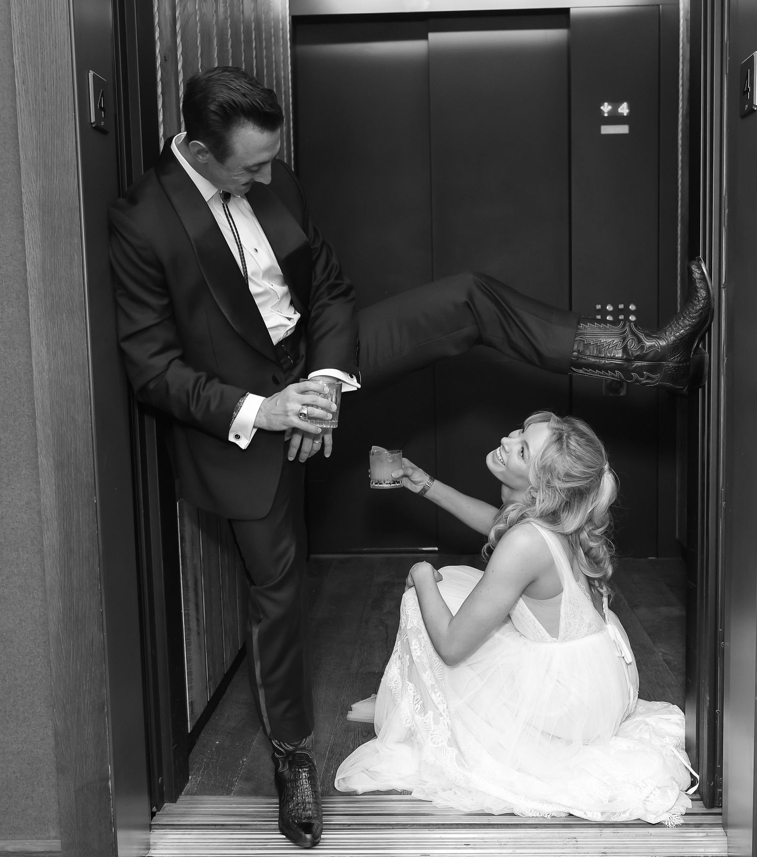 Black and white playful photo of bride sitting on elevator floor while groom holds her hand in formal attire.