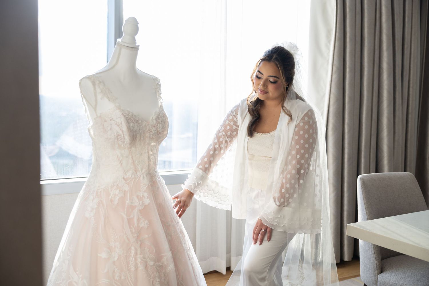 Bride in white robe admiring lace wedding dress on mannequin near bright window.