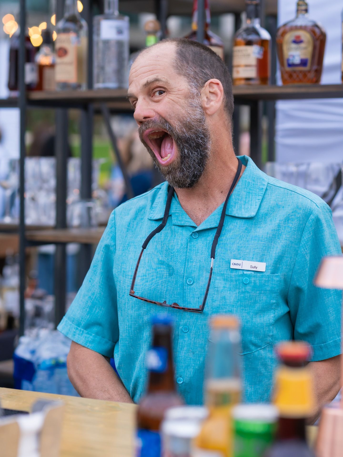 Expressive bartender in teal shirt reacting with surprise behind an outdoor bar at an event.