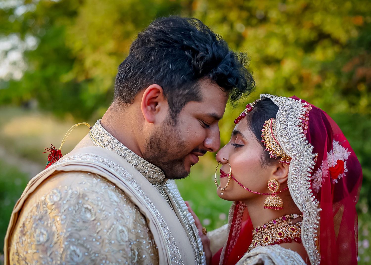 Indian bride and groom in traditional wedding attire share a romantic forehead touch outdoors.