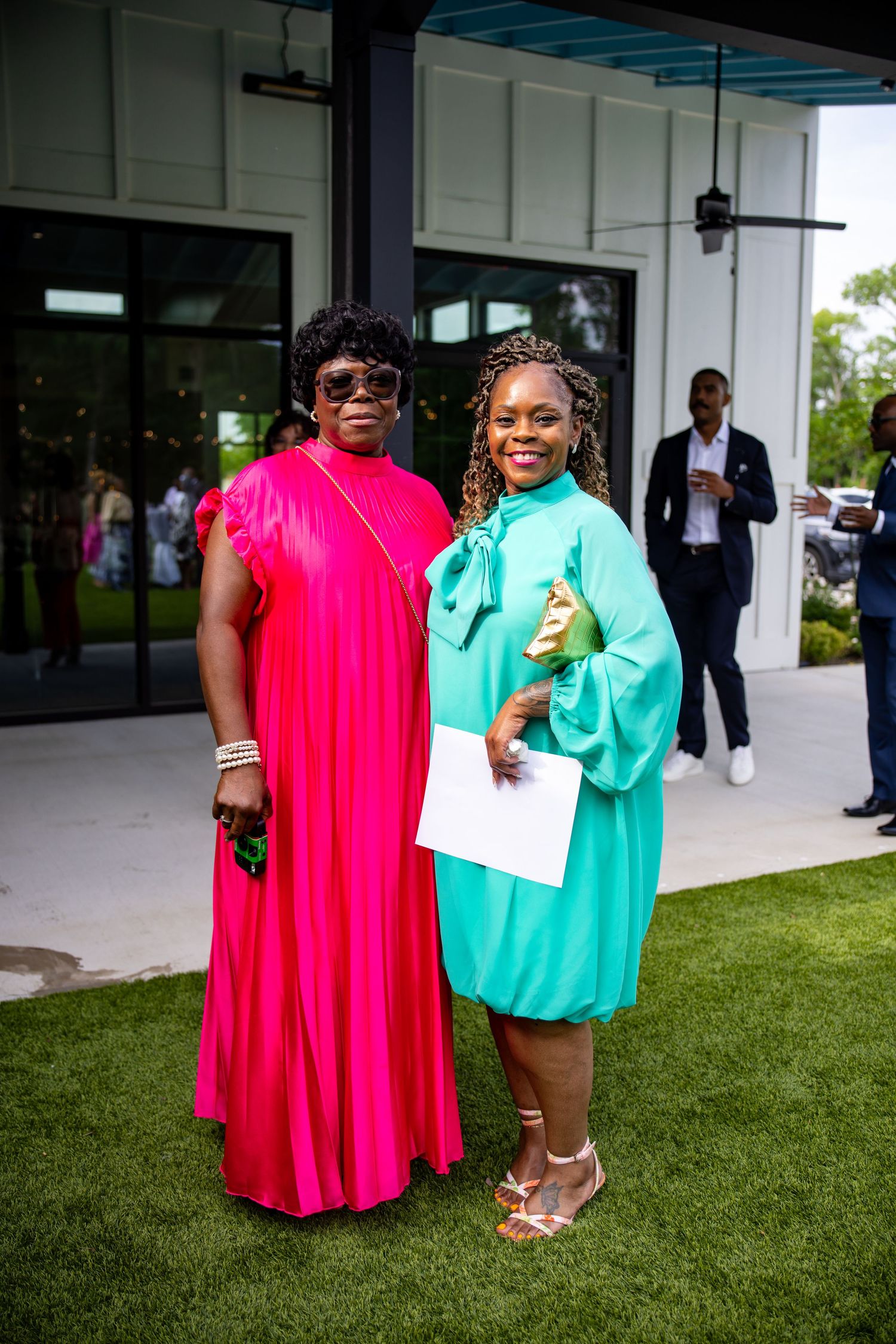 Two women posing outdoors in vibrant pink and teal dresses at a formal event on a green lawn.