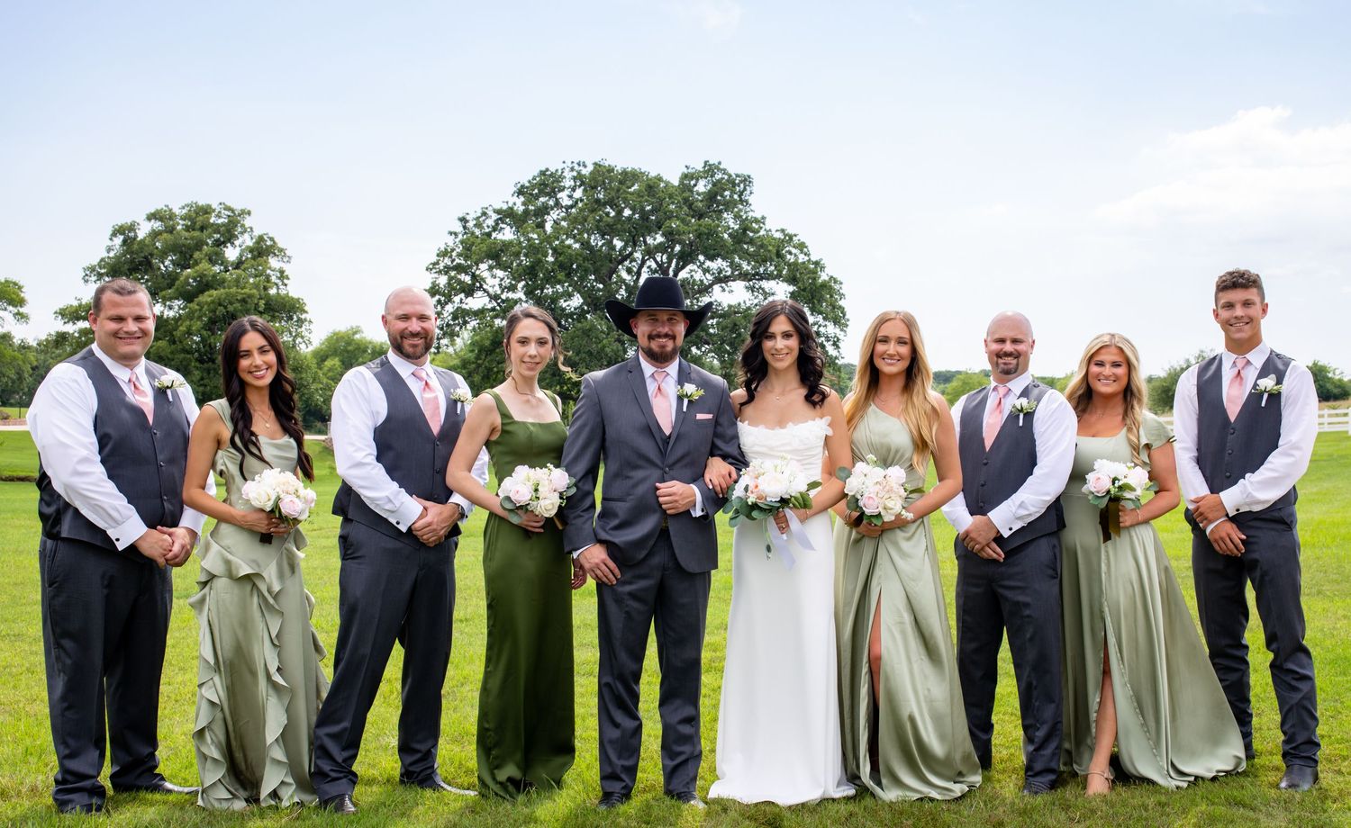 Wedding party poses outdoors in sage green and gray attire with a groom in a black cowboy hat on a lush green field.