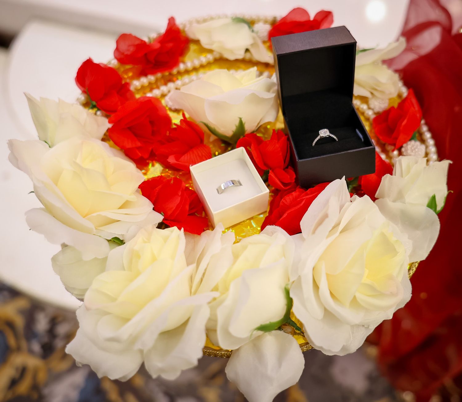 Two jewelry boxes with rings displayed on a decorative arrangement of white and red roses with pearl accents.