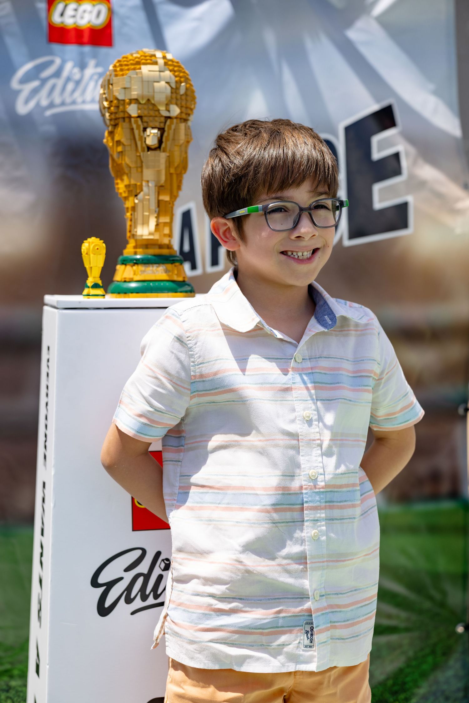Smiling boy with glasses poses beside a golden LEGO World Cup trophy at outdoor event.