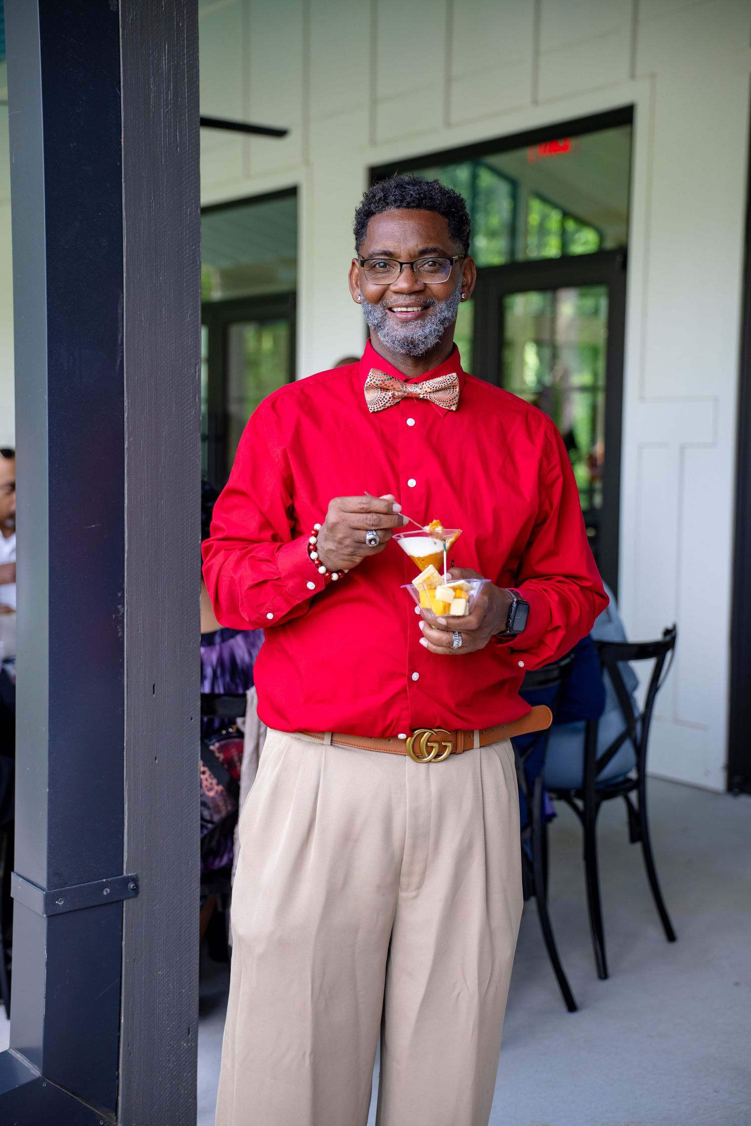 Smiling man in red shirt and bow tie holding a dessert cup outdoors at a restaurant patio.
