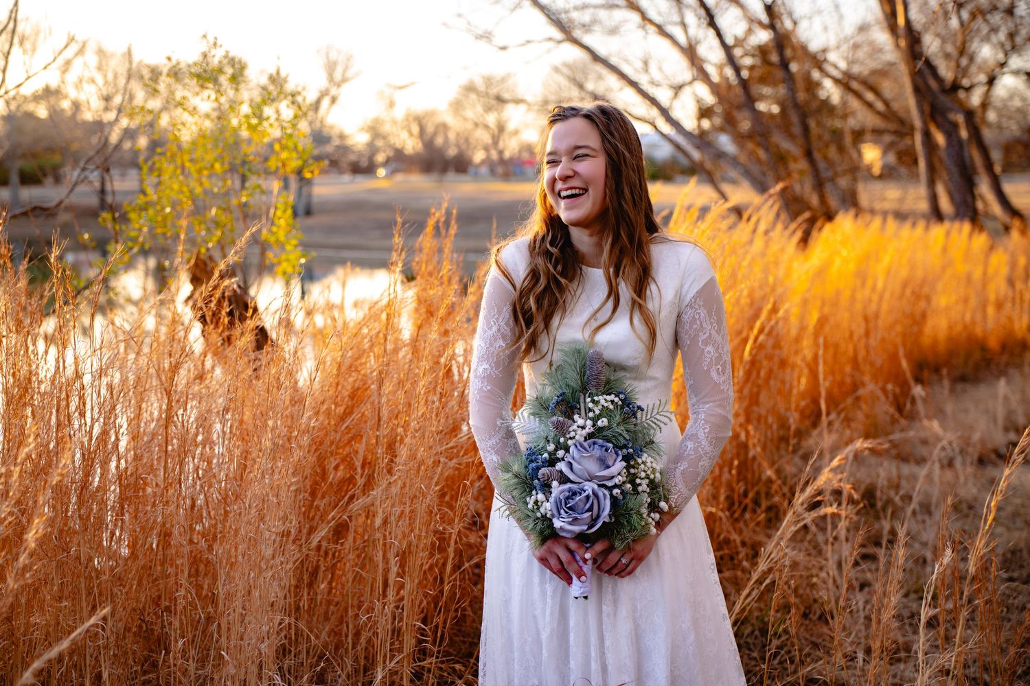 Joyful bride in white lace gown holding purple bouquet in golden autumn grass field.