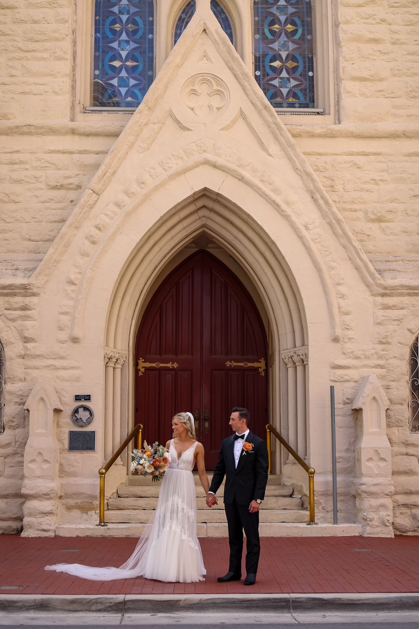 Bride and groom holding hands before ornate Gothic church entrance with stained glass windows and arched doors.