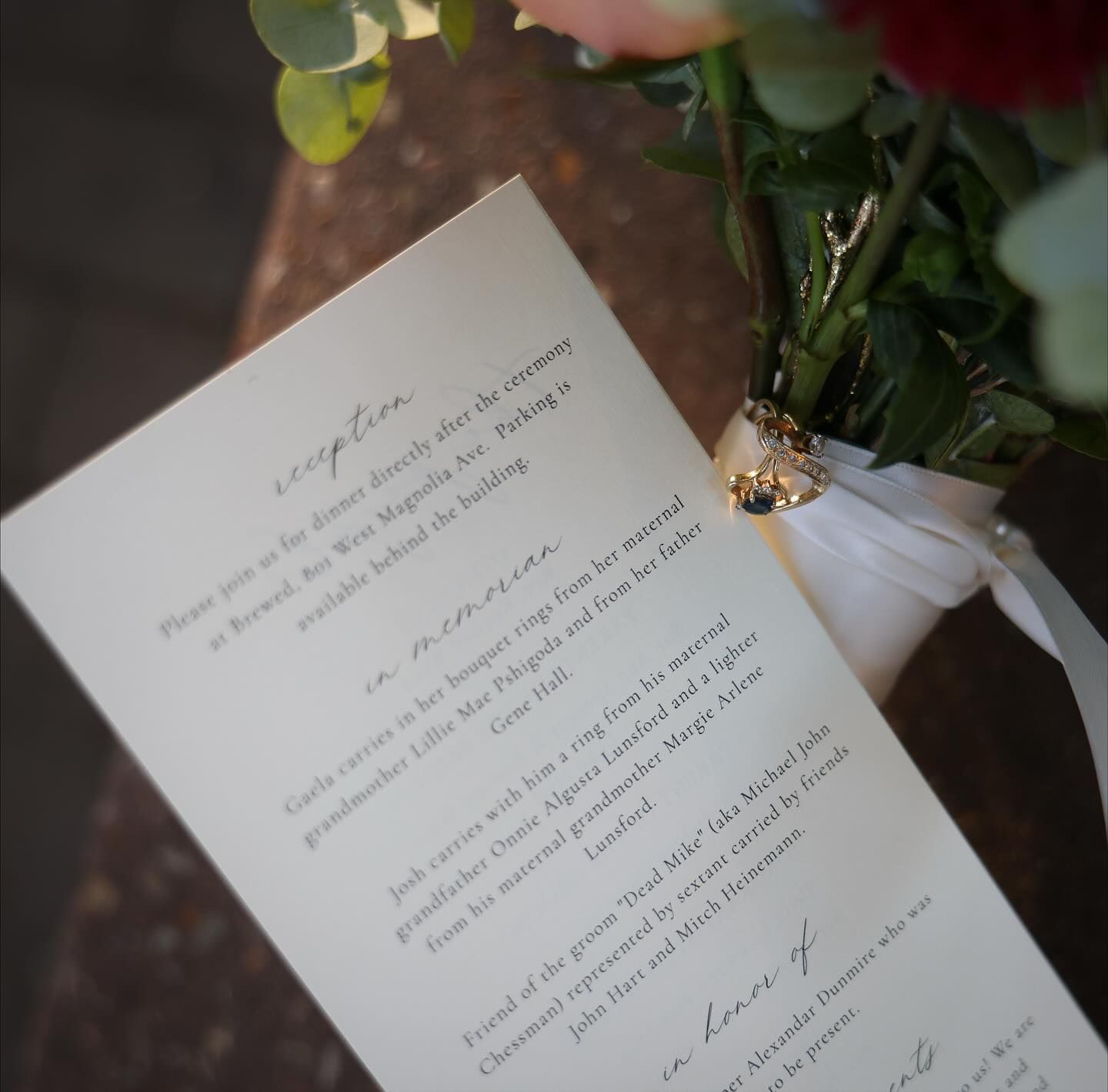 Elegant wedding program card with script text tied with white ribbon and pearl pin beside flowers.