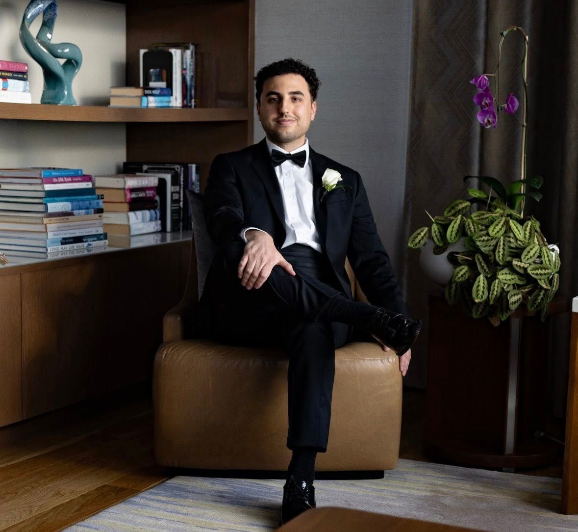 Dapper groom in black tuxedo with bow tie seated relaxed in leather chair before wedding ceremony.