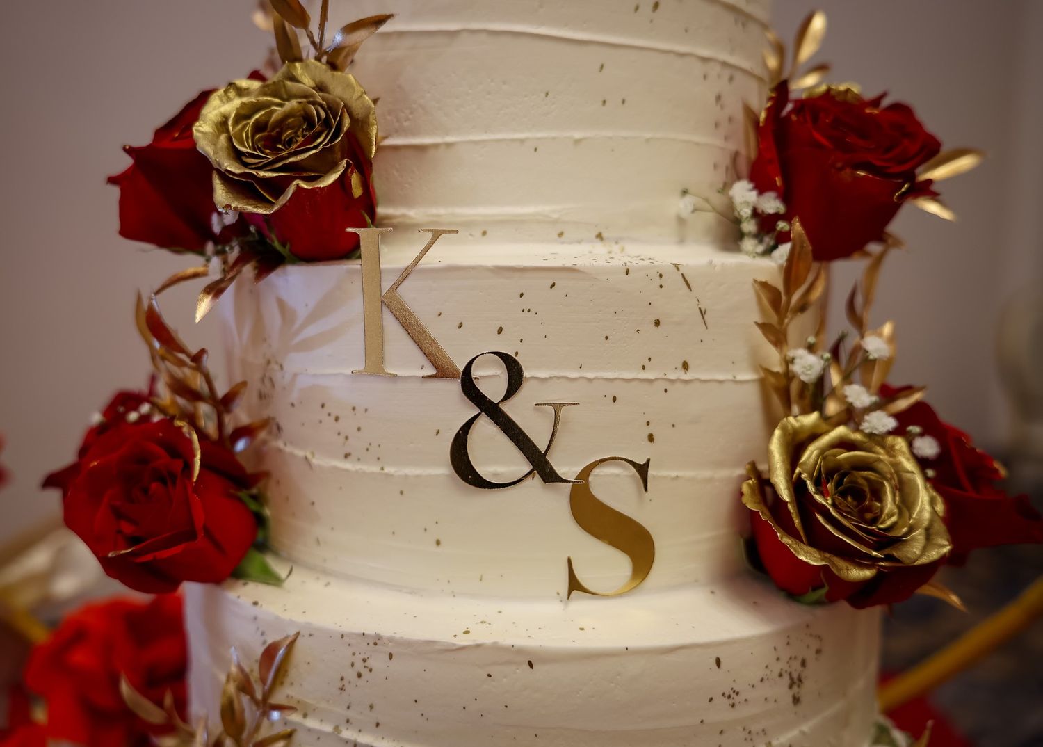 Three-tier white wedding cake with gold monogram K&S and red and gold roses.