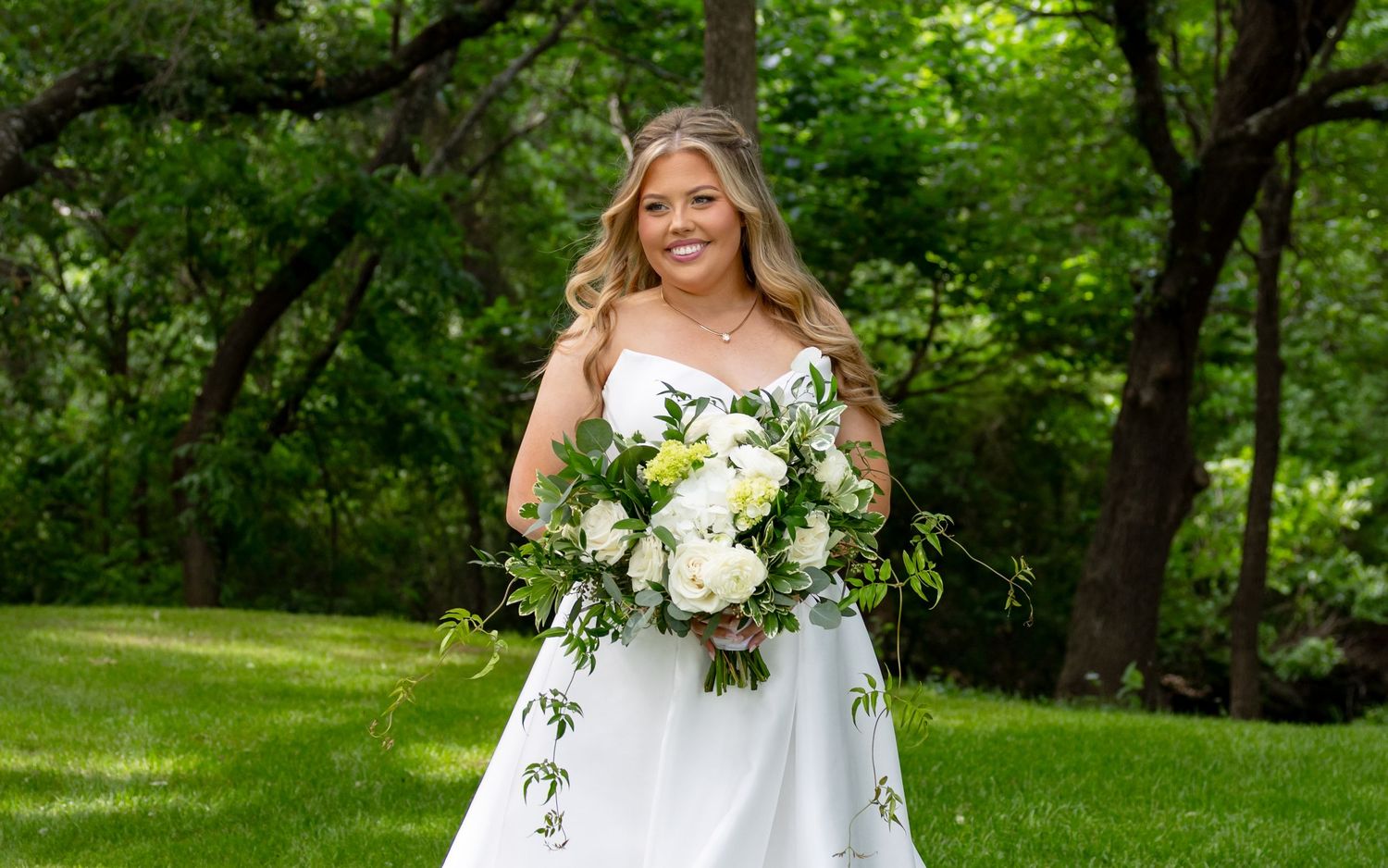 Smiling blonde bride in white gown holding white and green bouquet in lush garden setting.