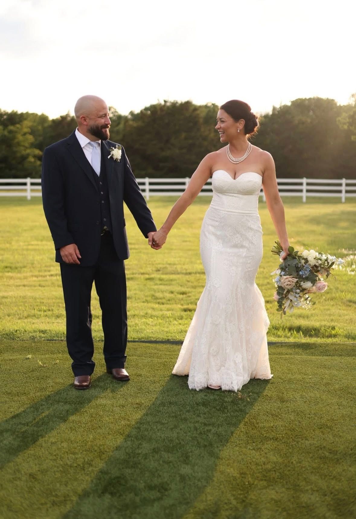 Bride in strapless mermaid gown and groom in navy suit holding hands on a sunlit green lawn at golden hour.
