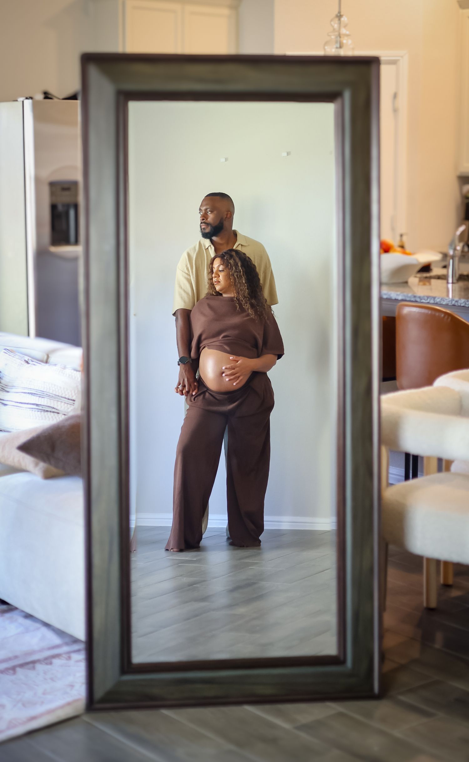 Couple poses in mirror reflection, pregnant woman in brown outfit cradled by partner in yellow shirt.