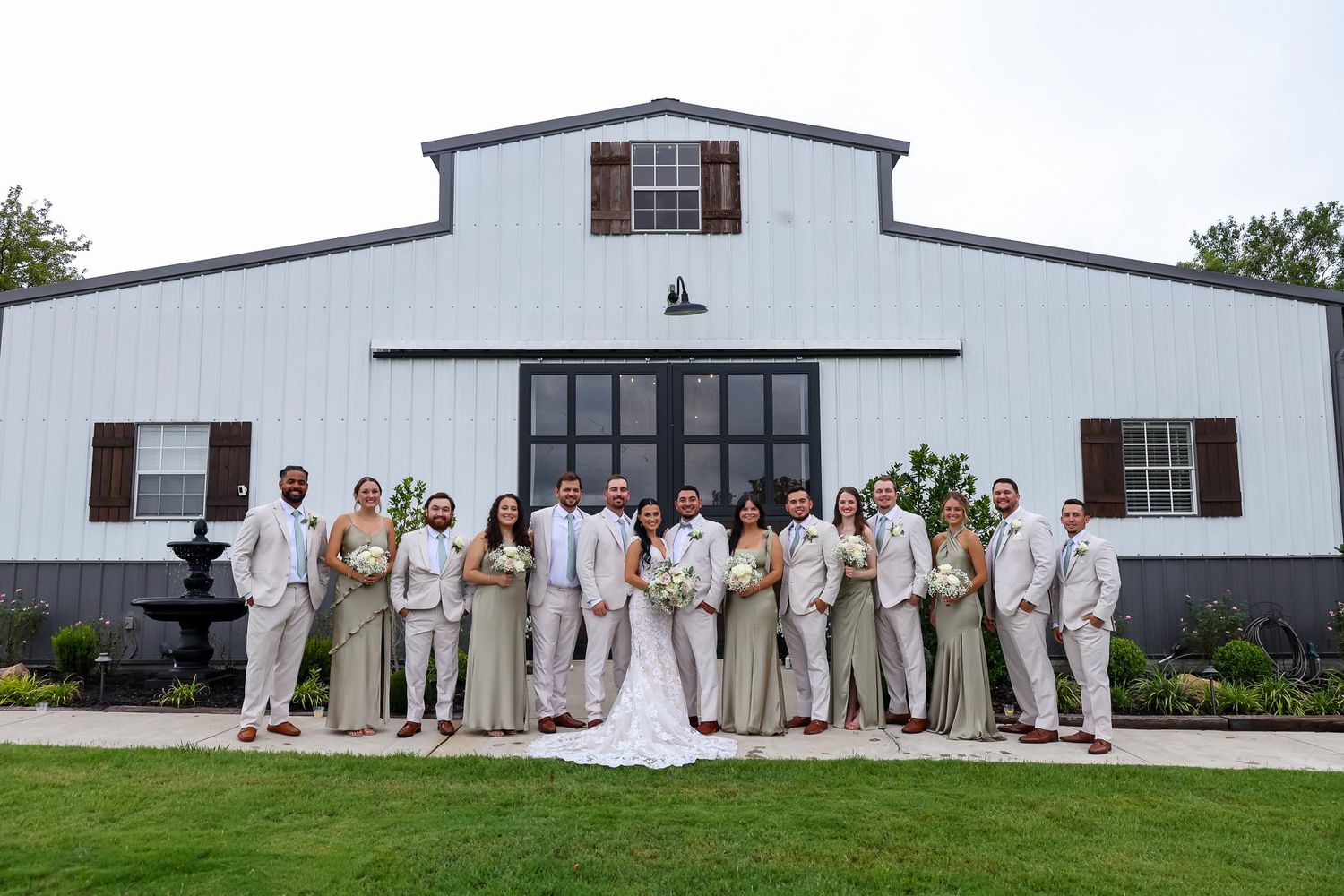Wedding party stands before a white barn venue, bridesmaids in sage dresses, groomsmen in tan suits.