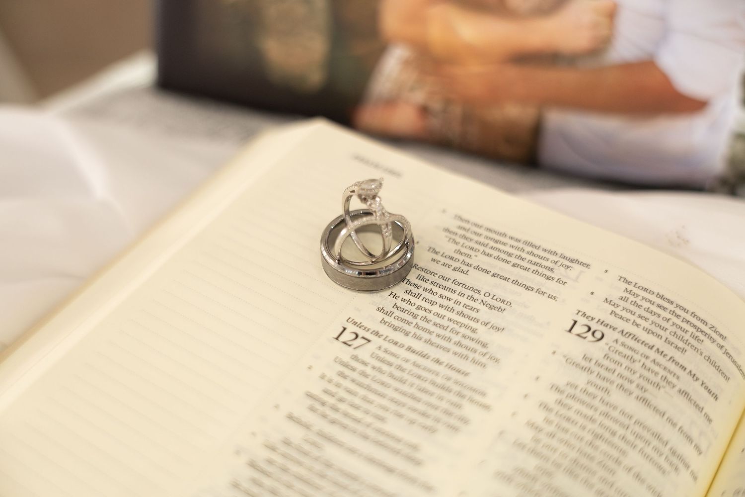Wedding rings resting on open Bible page with couple softly blurred in background.
