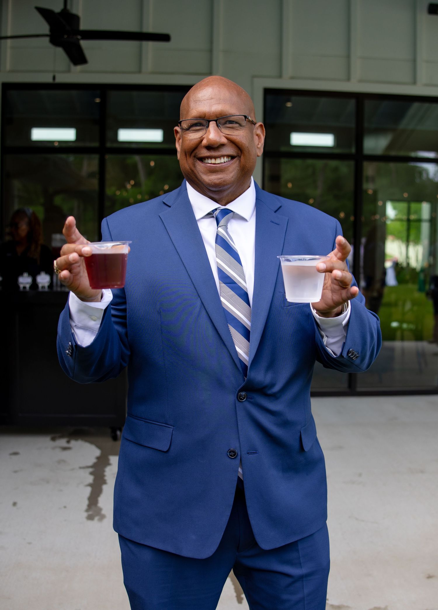 Man in blue suit smiling and holding two sample drinks outdoors at an event.