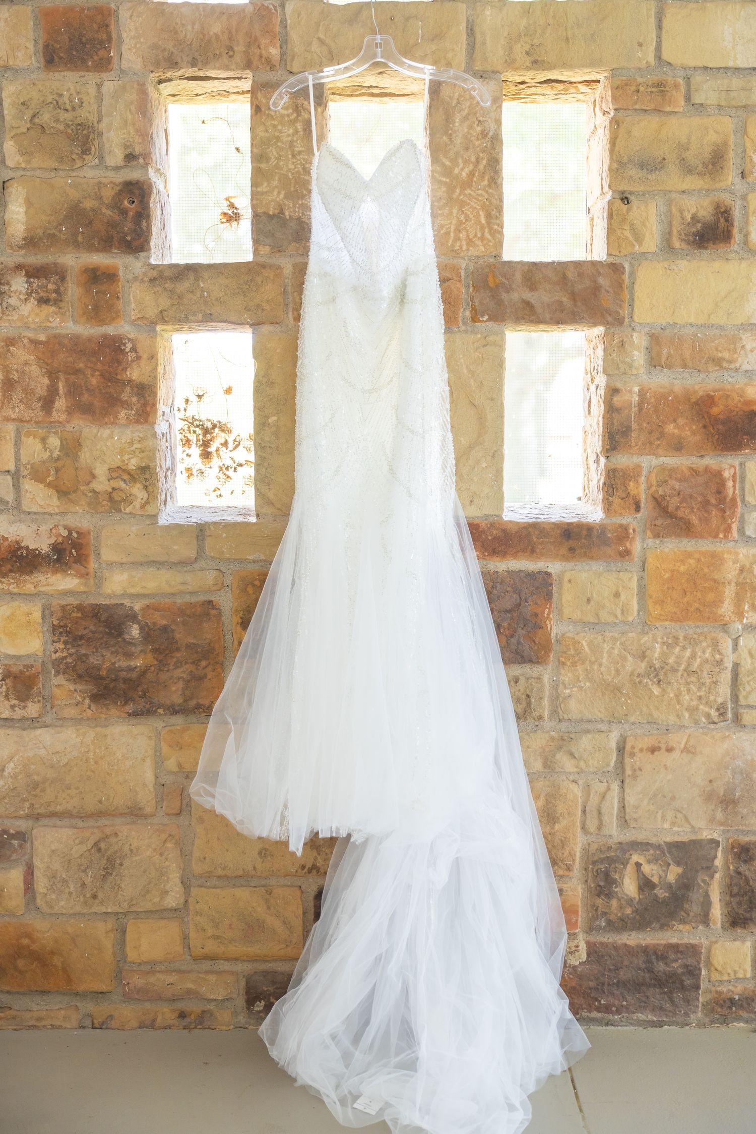 White strapless mermaid wedding gown with tulle train hanging against rustic stone wall.