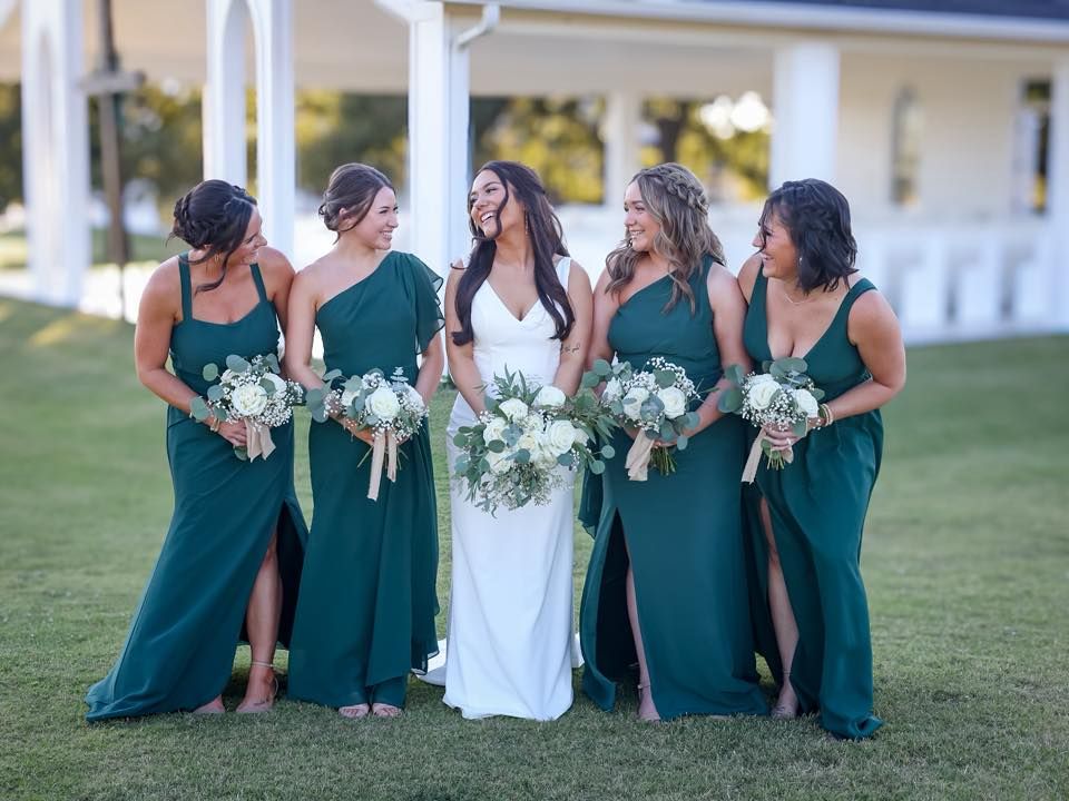 Bride with four bridesmaids in emerald green gowns holding white and greenery bouquets outdoors.