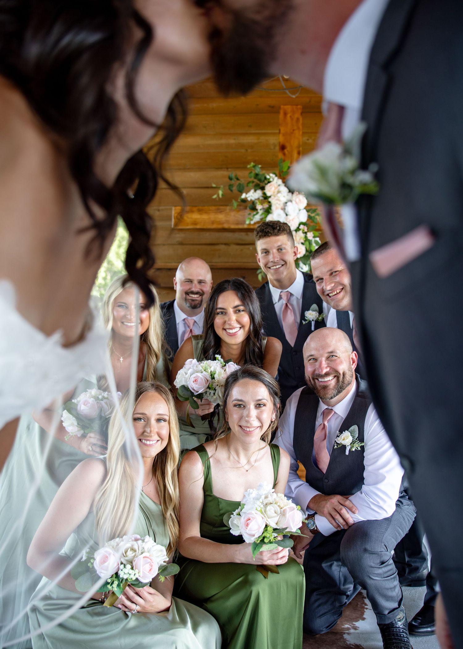 Wedding party smiles candidly as bride and groom stand in foreground at a rustic barn venue ceremony.