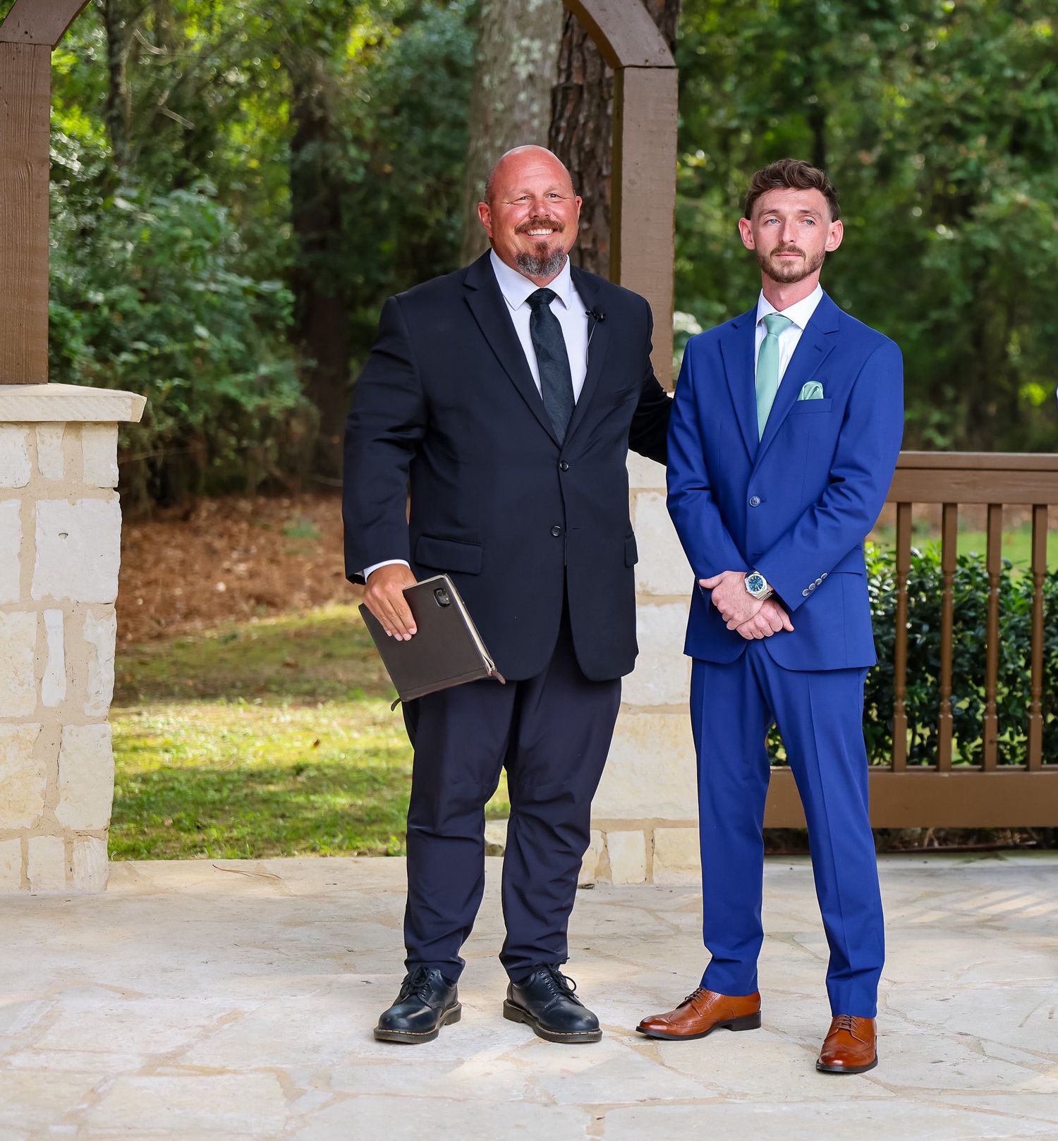 Groom in royal blue suit stands with officiant under stone arch at outdoor wedding venue.