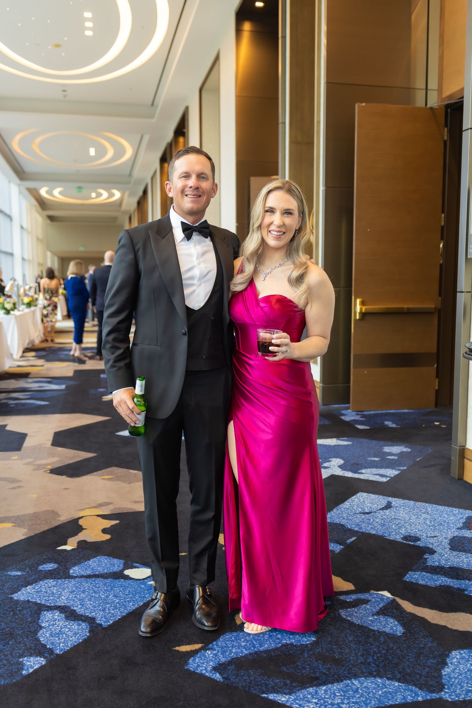 A couple in formal attire poses in a hotel hallway, woman wearing a vibrant pink gown.