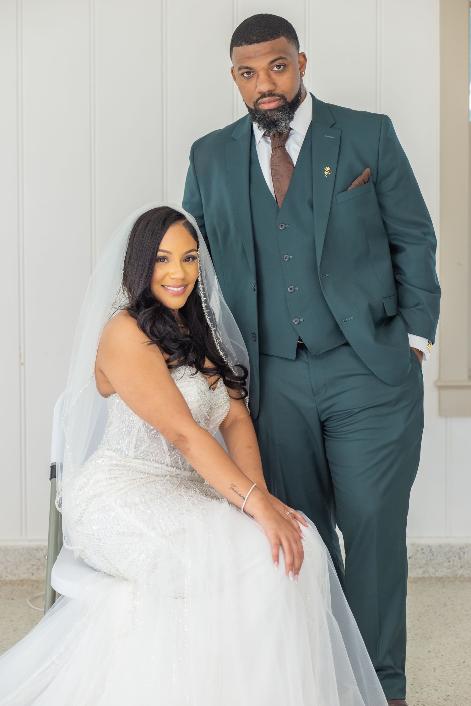 Bride in lace wedding gown seated while groom in emerald green suit stands beside her indoors.
