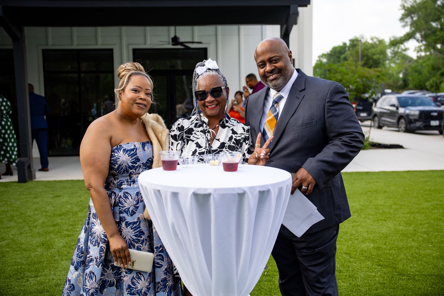 Three people smiling and posing together at an outdoor event around a cocktail table with drinks.