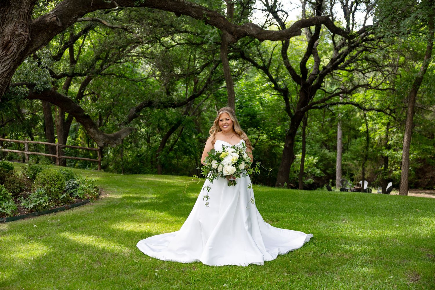 Beautiful bride in a white ball gown with long train poses in a lush green garden under large oak trees.