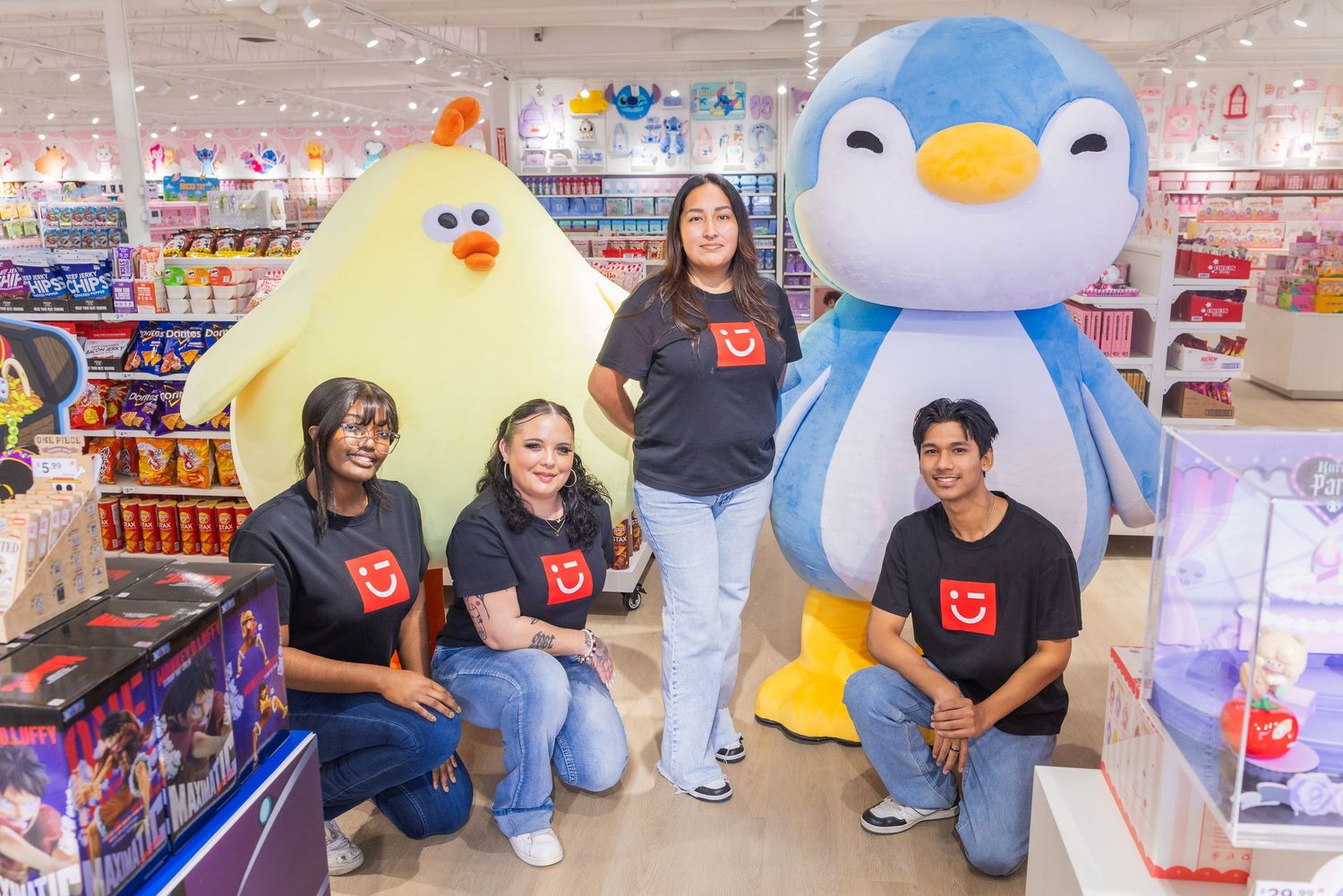 Four Miniso team members in branded black shirts posing with yellow chick and blue penguin mascots inside the store.