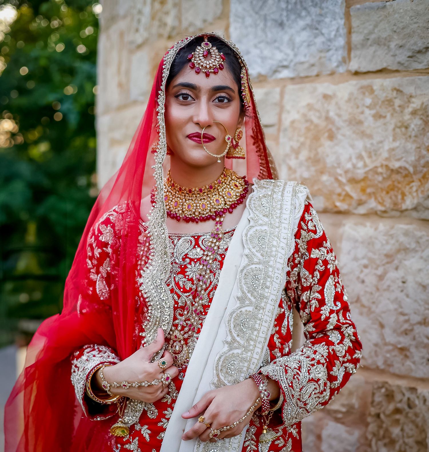 South Asian bride in red and gold bridal lehenga with traditional jewelry and red dupatta poses elegantly.