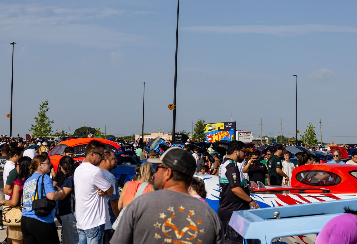 Large crowd gathered at an outdoor car show event with colorful classic cars on display under a blue sky.