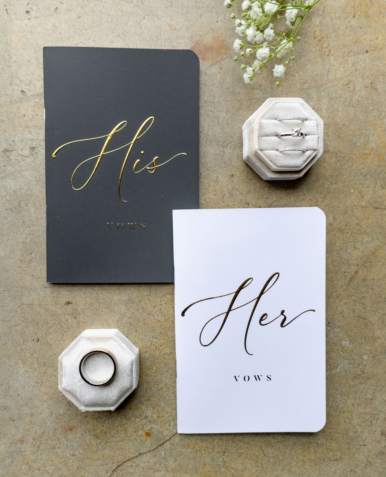 His and Her vow booklets with wedding rings in velvet octagon box and baby's breath flowers on stone surface.