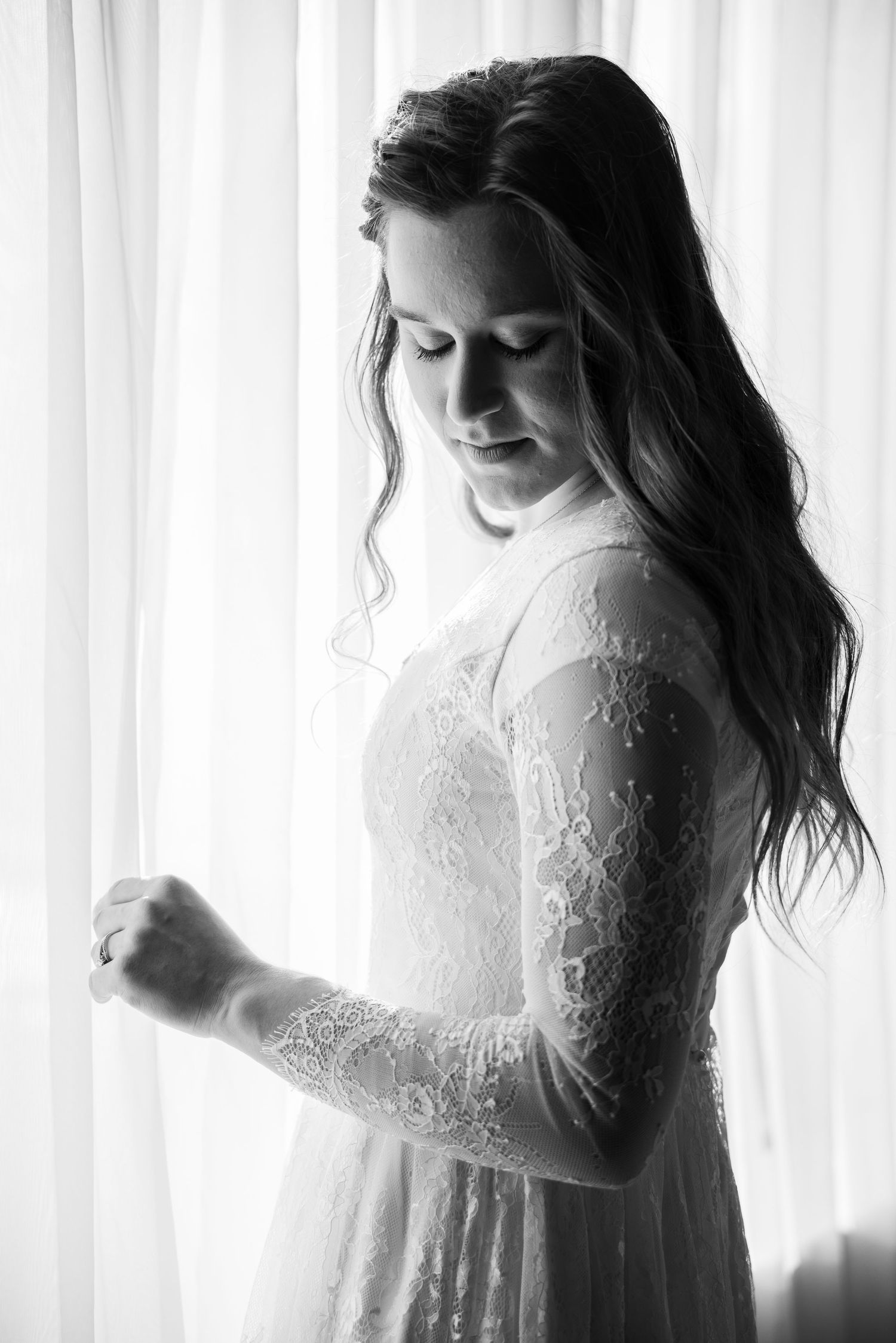 Black and white portrait of bride in lace wedding gown standing by curtained window.