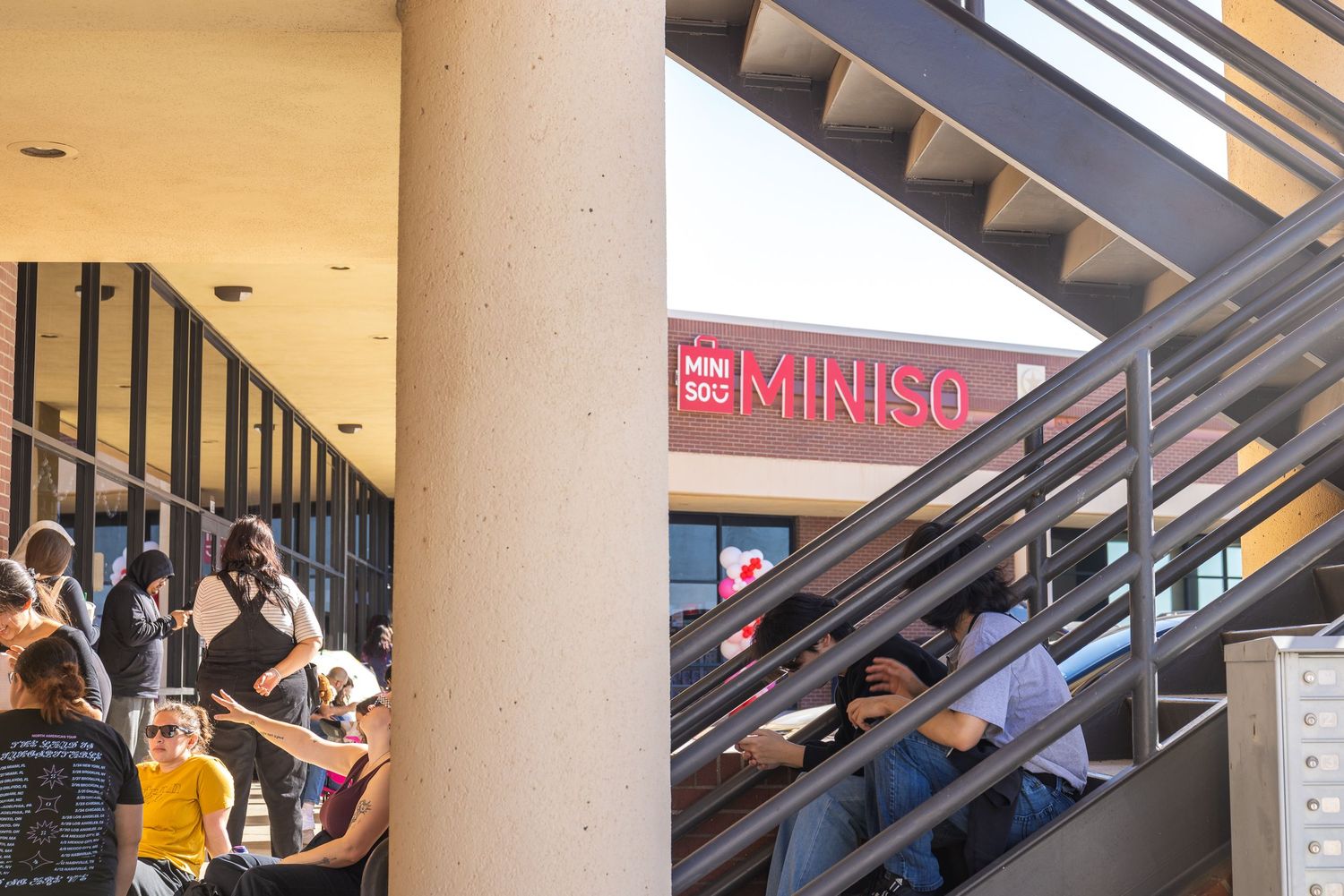 Exterior view of a MINISO retail store sign above a busy shopping mall entrance with escalators.