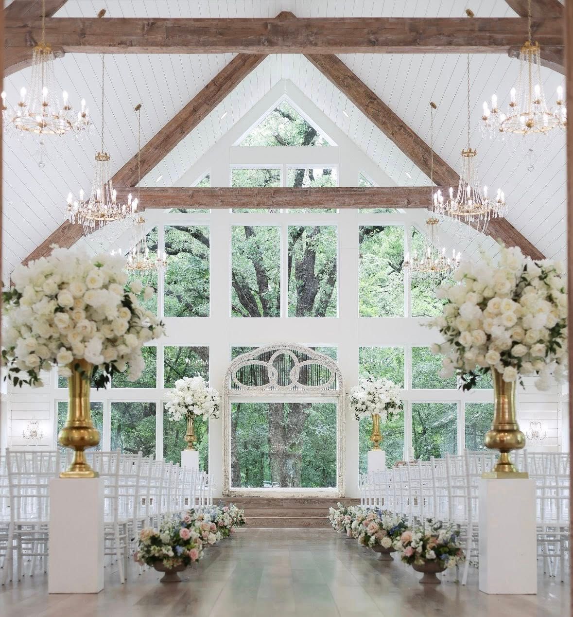 Elegant white wedding chapel with wooden beams, chandeliers, and tall white floral arrangements.