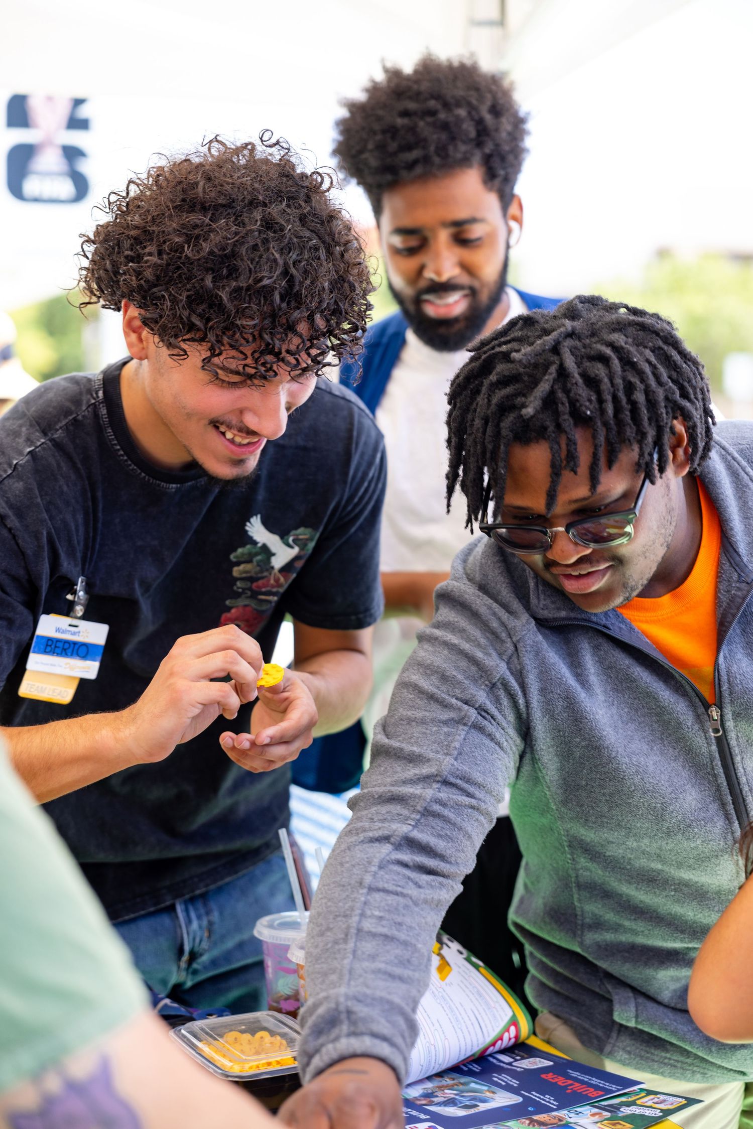 Three young men enthusiastically building with LEGO bricks at an outdoor community event.