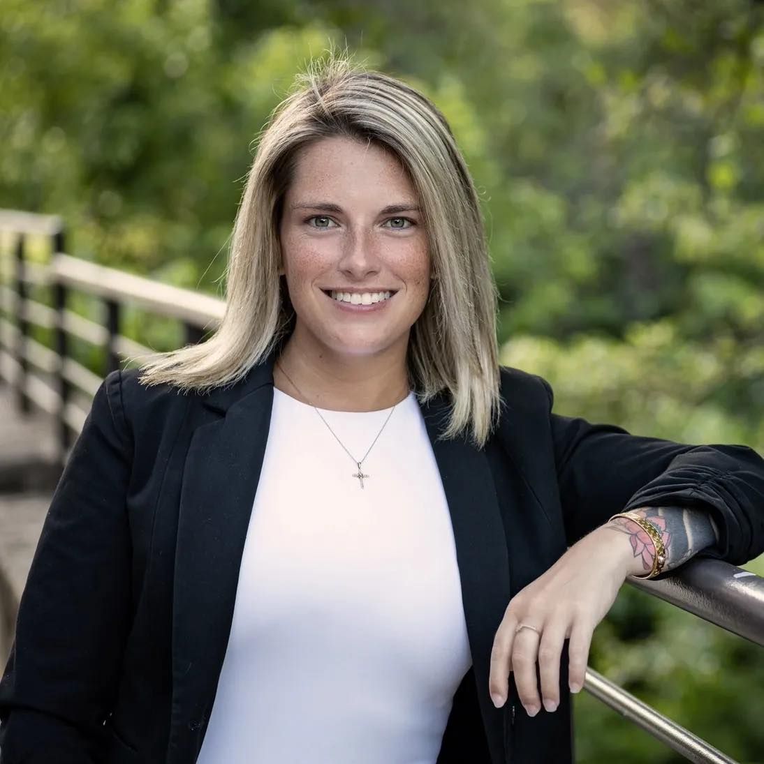 Smiling blonde woman in black blazer and white top poses outdoors near bridge with lush green trees background.