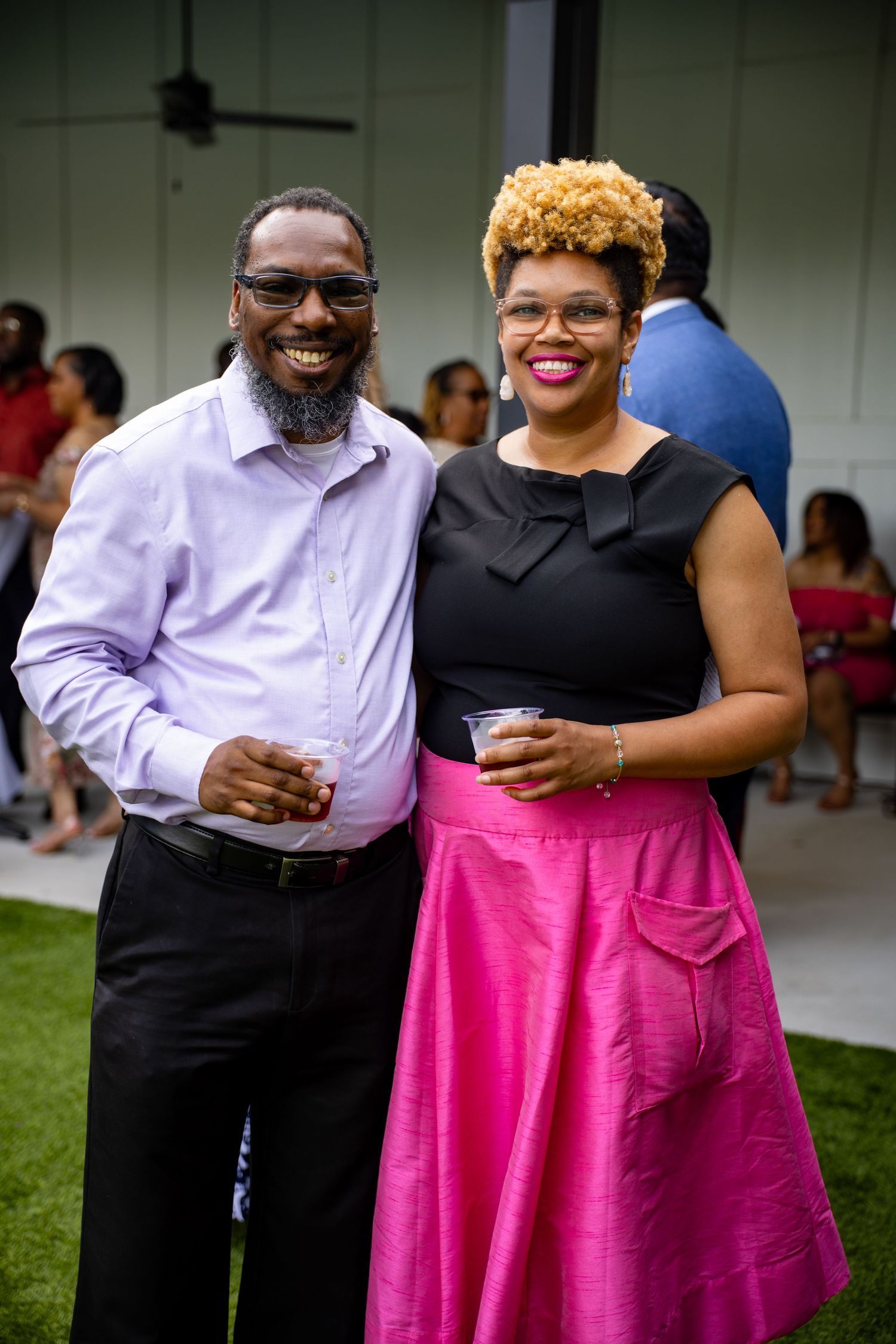 A smiling couple poses at an outdoor party, man in lavender shirt, woman in black top and pink skirt.