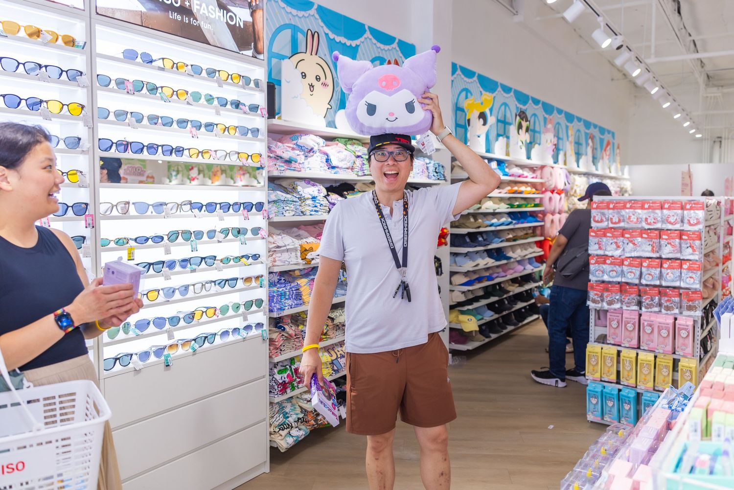 Customer trying on a large Kuromi plush hat while shopping in a Miniso store with sunglasses and toy displays nearby.