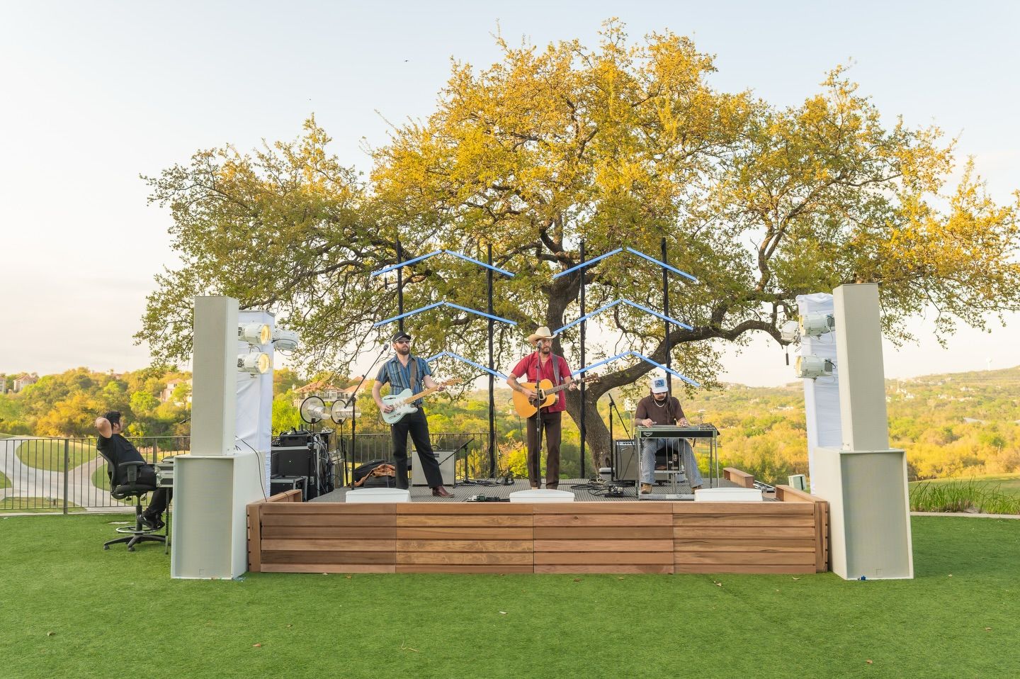 Live band performing on a wooden outdoor stage beneath a golden tree at sunset.
