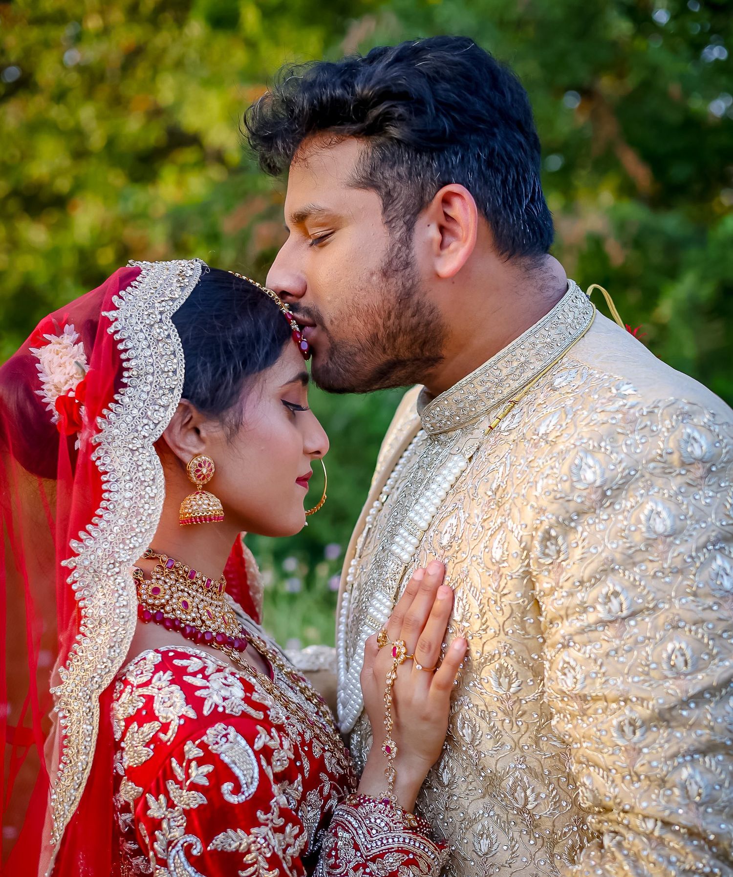 South Asian bride and groom in traditional red and gold wedding attire share an intimate moment outdoors.