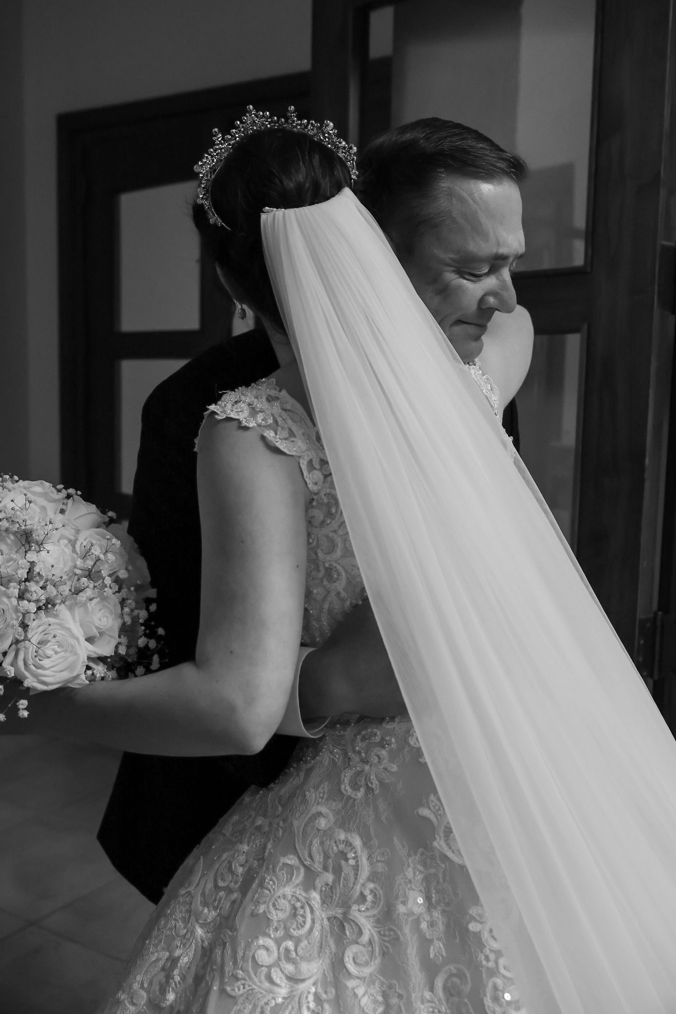 Black and white photo of bride and groom embracing, bride wearing lace gown, tiara, and veil holding bouquet.