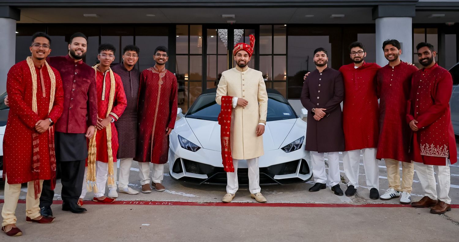 Groom in red sherwani poses with groomsmen in traditional attire beside a white Lamborghini.