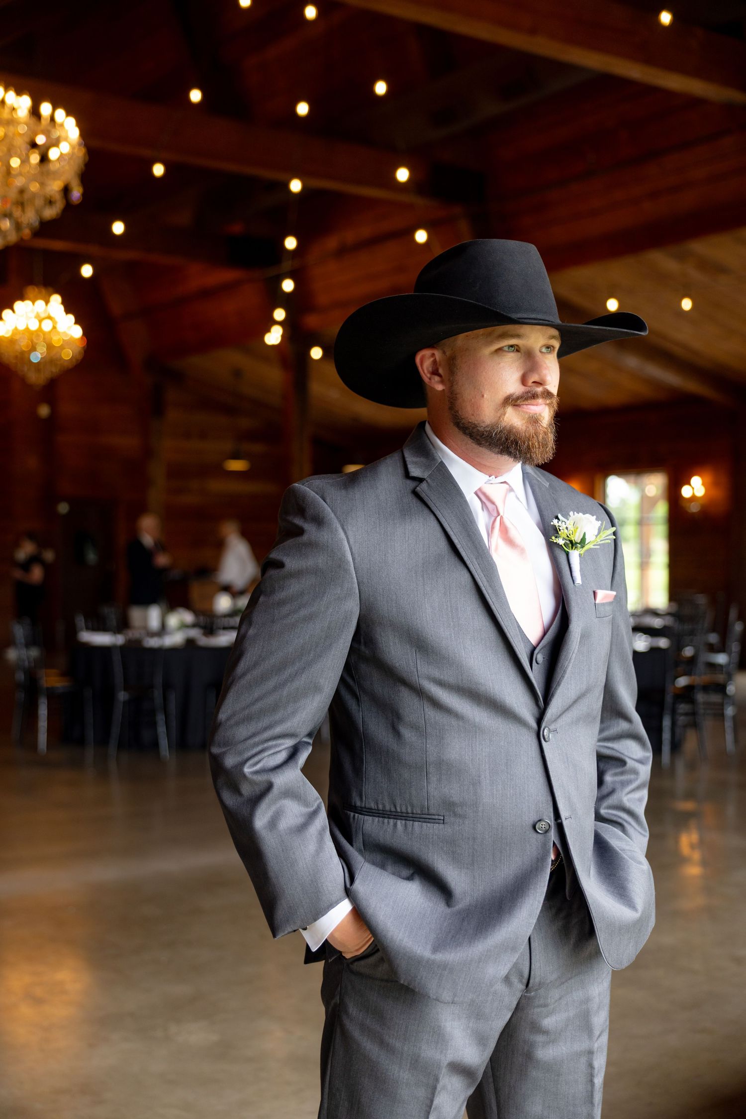 Handsome groom in gray suit, black cowboy hat, and boutonniere stands confidently in rustic barn venue.