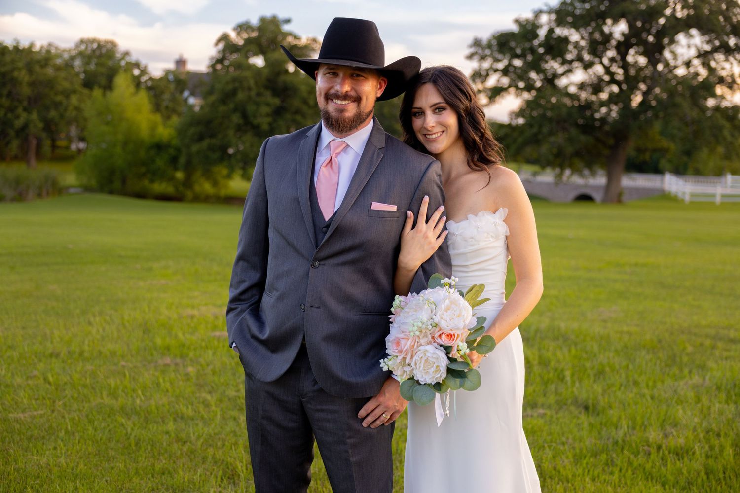 Happy newlyweds pose outdoors on green grass, groom wearing black cowboy hat and gray suit with pink tie.