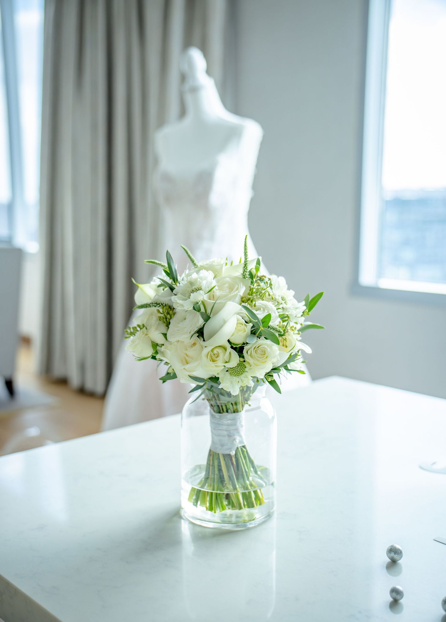 White bridal bouquet of roses and tulips in glass vase with wedding dress on mannequin behind.