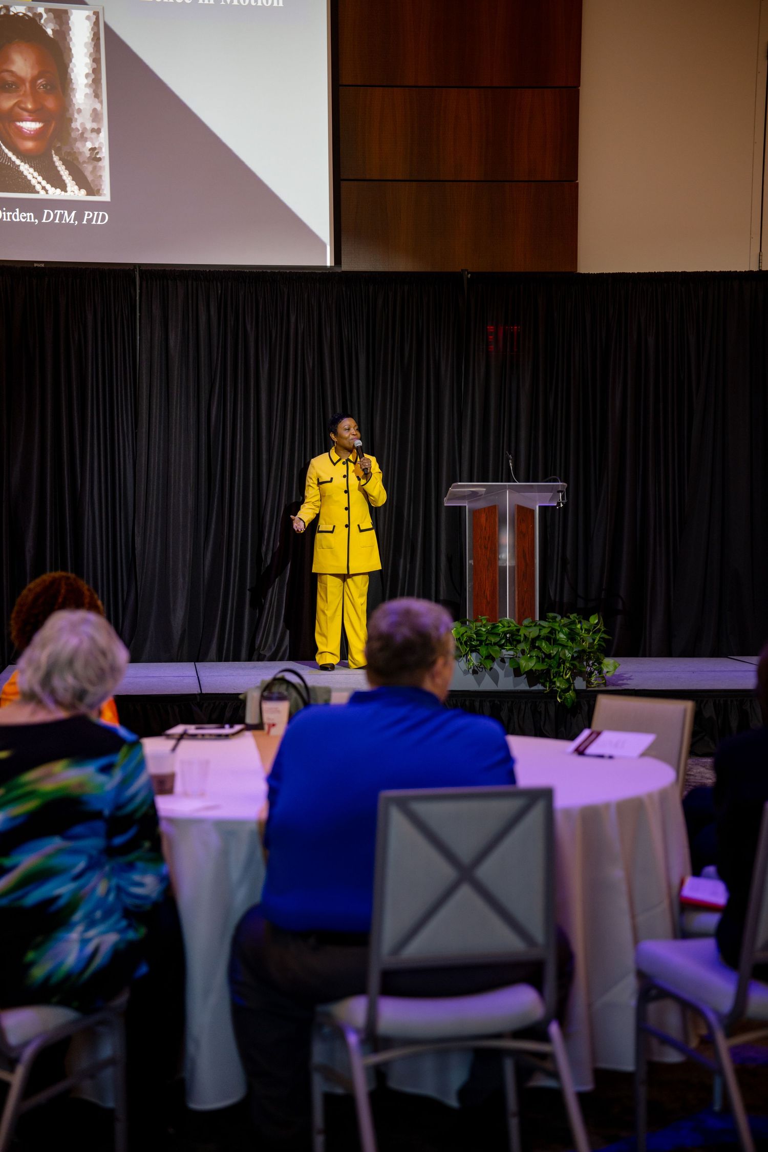 A woman in a yellow pantsuit speaks on stage to seated attendees at a banquet-style conference event.