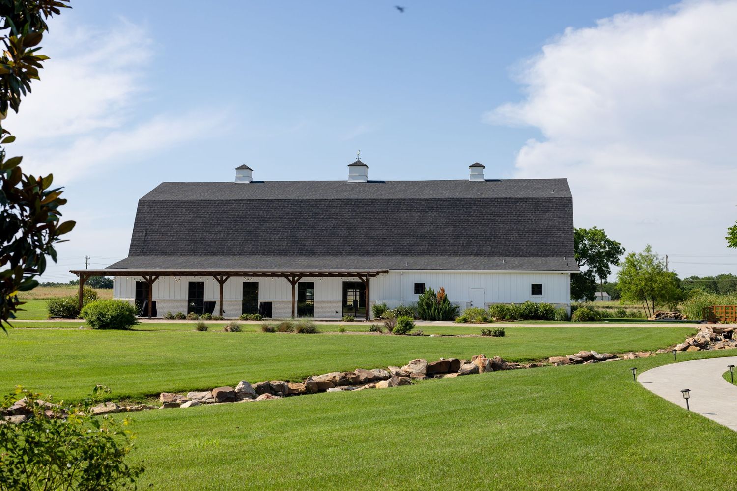 White barn wedding venue with gray roof sits on manicured green grounds under a bright blue sky.