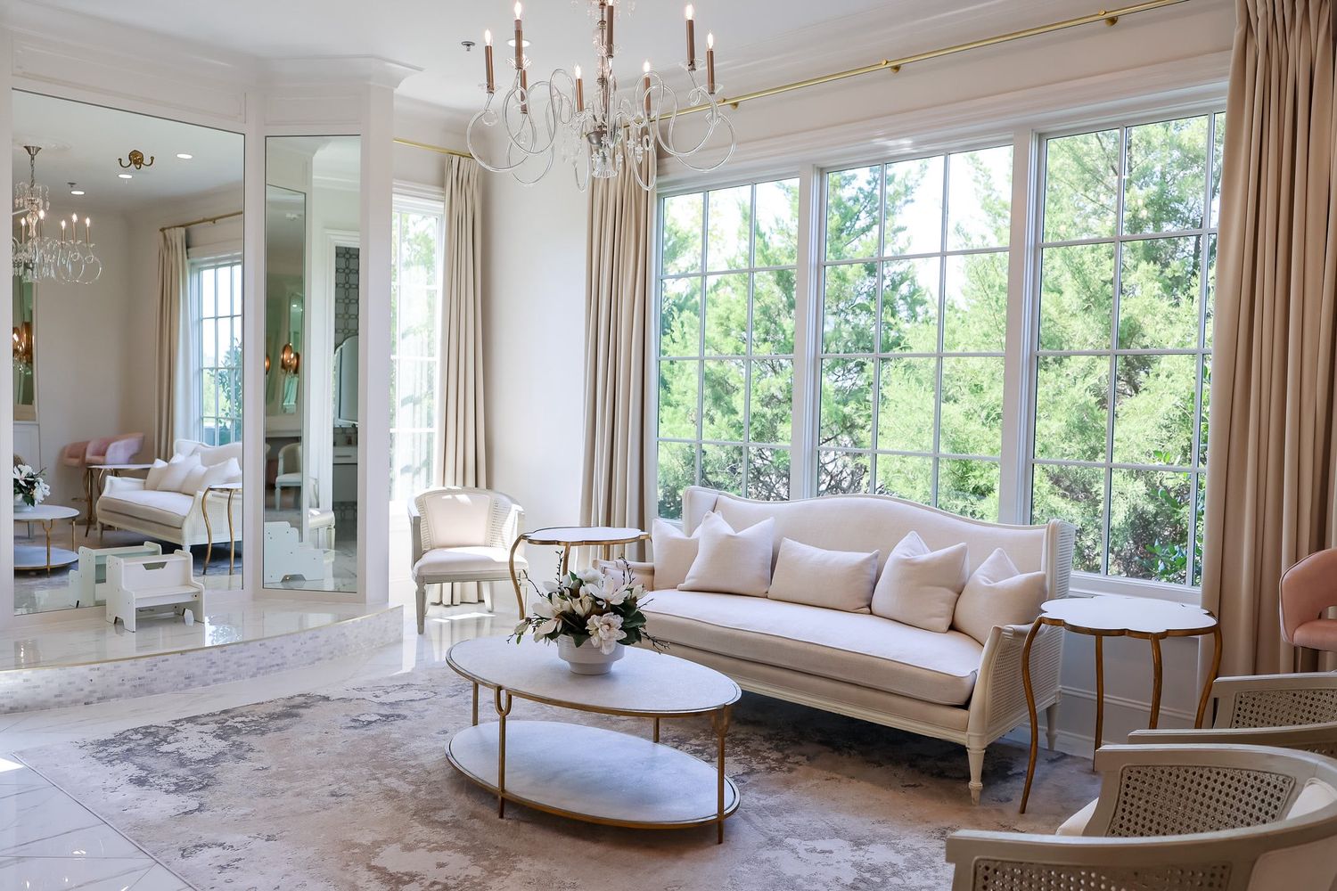 Luxurious white living room with chandelier, elegant sofa, gold accents, and large windows.